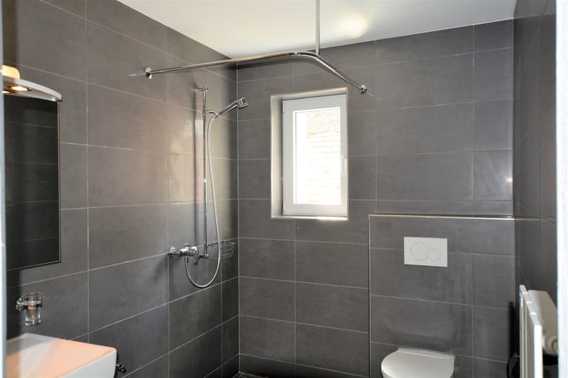 Spacious bathroom with gray tile walls, a glass-enclosed shower with a chrome shower head, and a small window providing natural light.