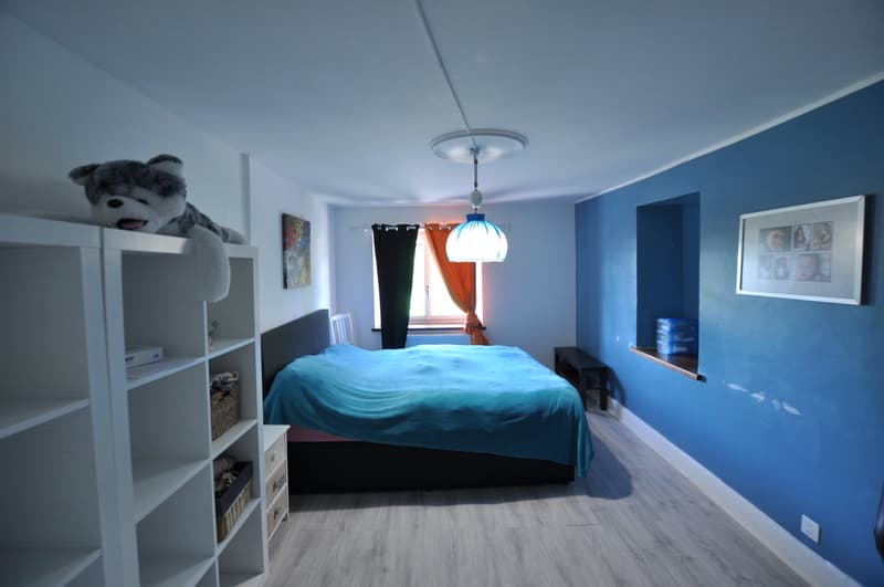 Spacious bedroom with blue walls, hardwood flooring, a large bed with a teal comforter, a shelving unit, and a window with curtains.