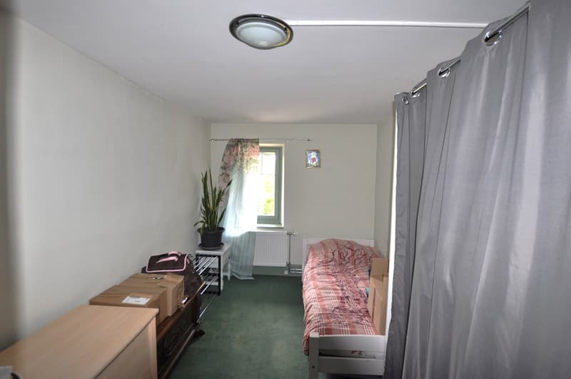 The image shows a bedroom with a bed, dresser, and storage boxes. The room has white walls, a green carpet, and a curtain covering the window. There is also a potted plant and a framed picture on the wall.