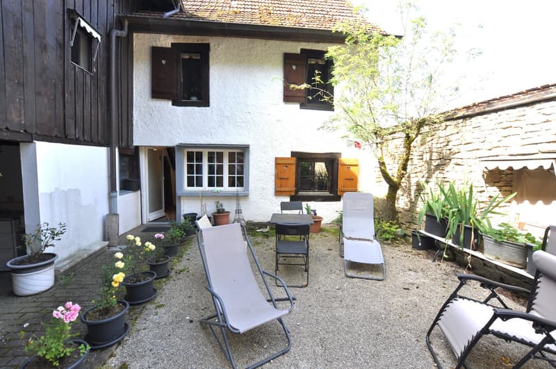 Two-story house with white exterior walls, wooden shutters, and a gravel courtyard. The courtyard contains potted plants, outdoor furniture, and a small tree. The house appears to have a balcony or terrace on the upper level.