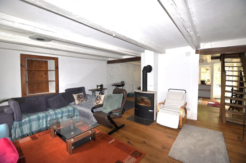 Cozy living room with hardwood floors, white walls, exposed wooden beams, a sofa with a plaid blanket, a coffee table, a recliner chair, and a wood-burning stove fireplace.