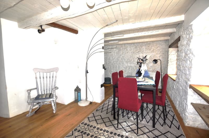 Rustic dining room with exposed wooden beams, stone wall, red upholstered chairs, wooden table, and a rocking chair in the corner