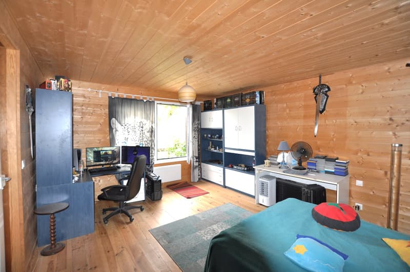 Wooden paneled walls and ceiling, desk with computer setup, shelving unit, bed, and various decor items