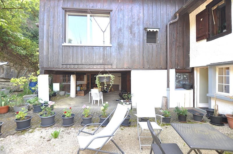 Rustic wooden house with a balcony, surrounded by potted plants and outdoor furniture. The house has a cozy, homely atmosphere with a charming, natural setting.
