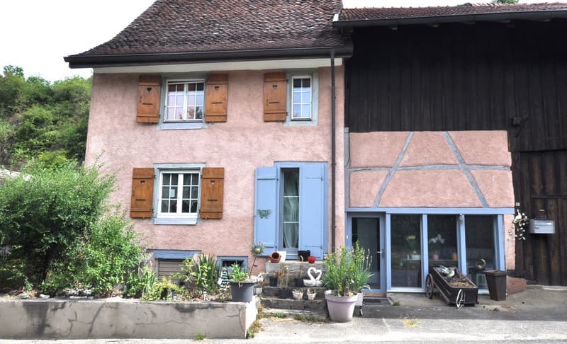 Two-story house with a pink exterior, wooden shutters, and a tiled roof. The house has a balcony or terrace on the upper level. There are potted plants and decorative elements around the entrance.