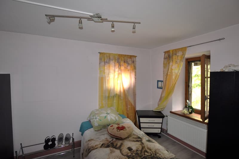 Bedroom with white walls, hardwood floor, large window, yellow curtains, dresser, and a bed with a fur blanket and decorative pillows.