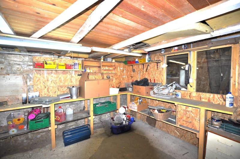 The image shows a cluttered basement or storage room with wooden shelves and storage containers. The room has a wooden ceiling with exposed beams and a concrete floor. Various household items, tools, and other miscellaneous objects are stored on the shelv