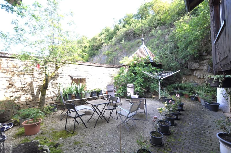 Cozy outdoor patio area with stone walls, wooden roof, potted plants, and a table with chairs for relaxing and dining. The patio is surrounded by lush greenery and trees, creating a peaceful and secluded atmosphere.