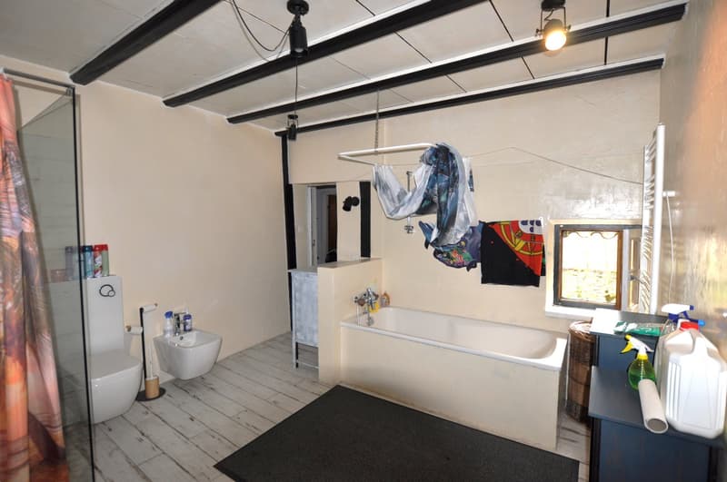 Bathroom with a bathtub, toilet, and a washing machine. The room has exposed wooden beams on the ceiling and a window. There are various cleaning supplies and personal items visible.