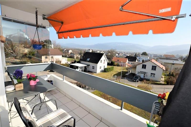 Balcony with table, chairs, and awning, surrounded by tile floor, overlooking a grassy area with distant houses