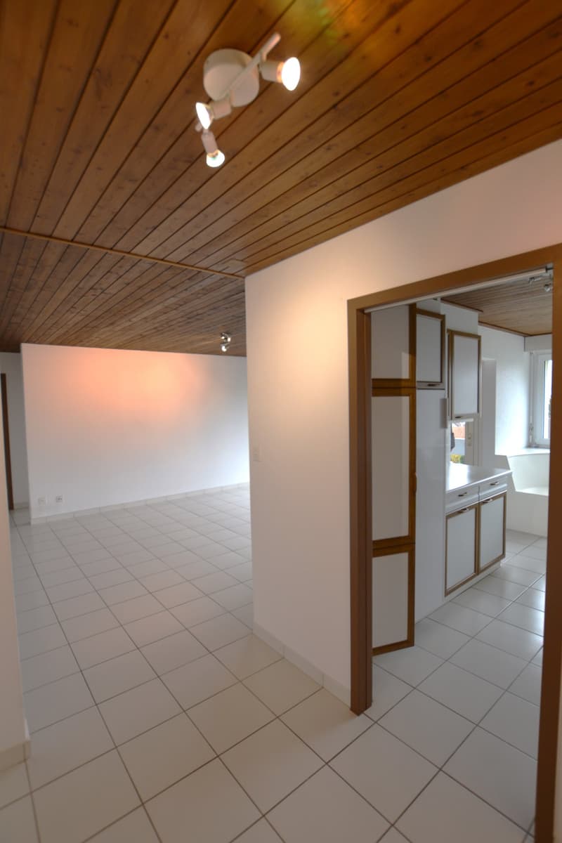 Empty room with white tile flooring, wooden ceiling, white walls, kitchen area with cabinets and a window