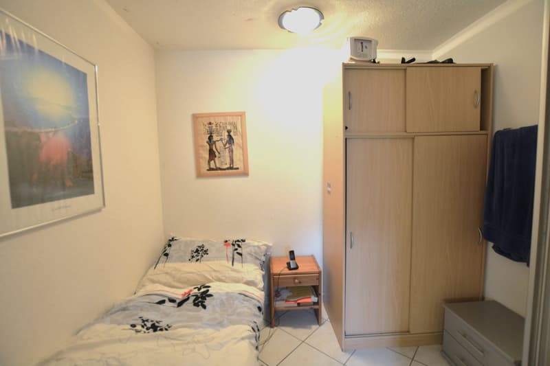 Single bed with patterned bedding, a nightstand with a cordless phone, a picture frame, and a wooden wardrobe. A small table with a towel and some towels on it.
