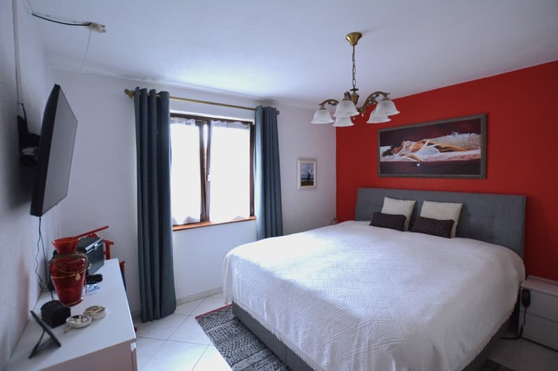Bedroom with a large bed, red wall, white tile floor, a cabinet with various items on top, mounted television, window with white and blue curtains, and a picture frame hanging on the wall
