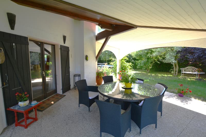 terraced covered outdoor dining area with table and chairs, small table with flower vase, house door, green plants, green lawn, white metal bench