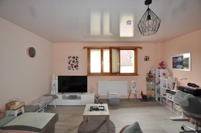 Large living room with sofa, desk, TV, rug, radiator, desk lamp, window, shelving, chair, clock, and art on the wall