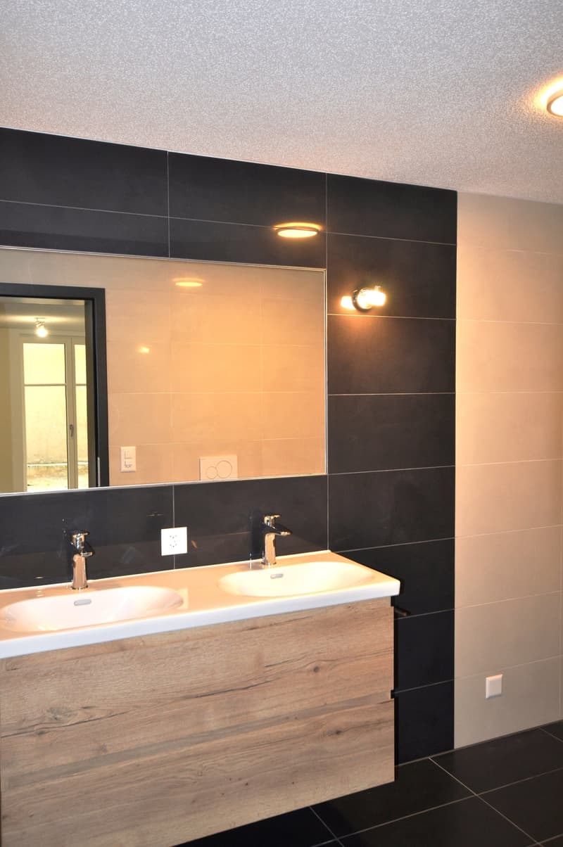 A modern bathroom with double sinks, black tiles, a wooden cabinet, and a large mirror. Faucets are silver, power outlets are white.