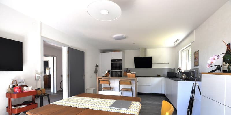 A modern kitchen with a dining area, white cabinets, tiled countertops, and an open floor plan. A wooden table with a laptop and a yellow chair in the center of the room.