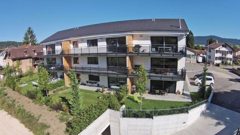 A three story white building with multiple balconies, green lawn, and a gravel parking lot