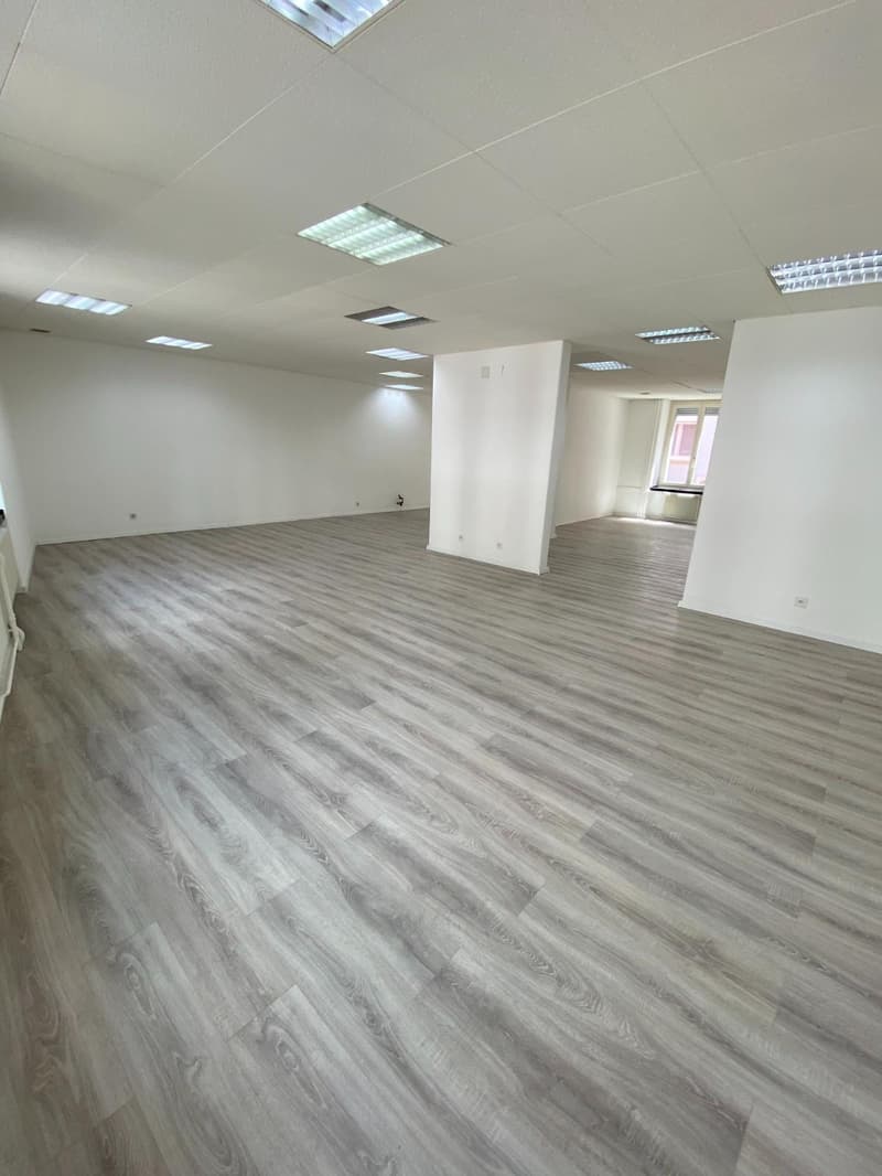 large empty room, grey wooden floor, white walls, white ceiling with recessed lighting, power outlets on the wall, window