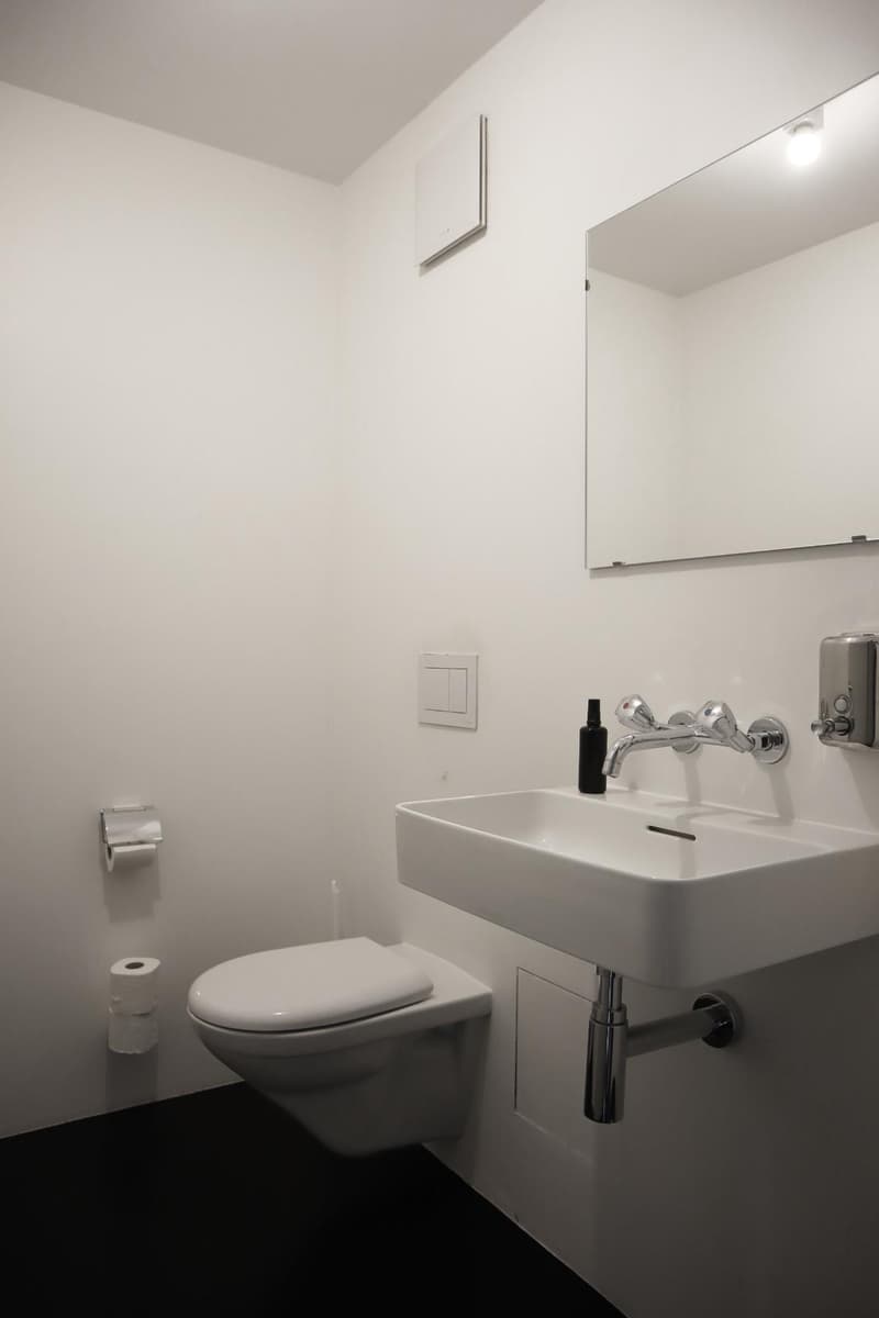 White tiled walls, white sink with a silver faucet, white toilet, a mirror, wall-mounted toilet paper holder, and a soap dispenser.