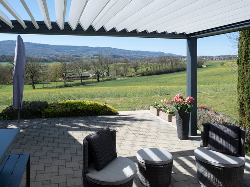 Outdoor seating area with a view of a green field, featuring two wicker chairs, a table, and a closed umbrella.