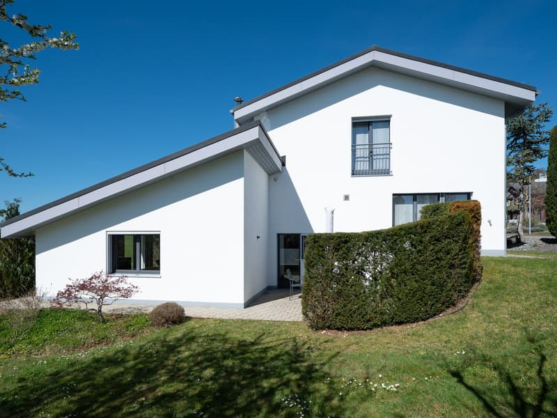 Modern white house with a flat roof, large windows, and a small garden with a hedge.