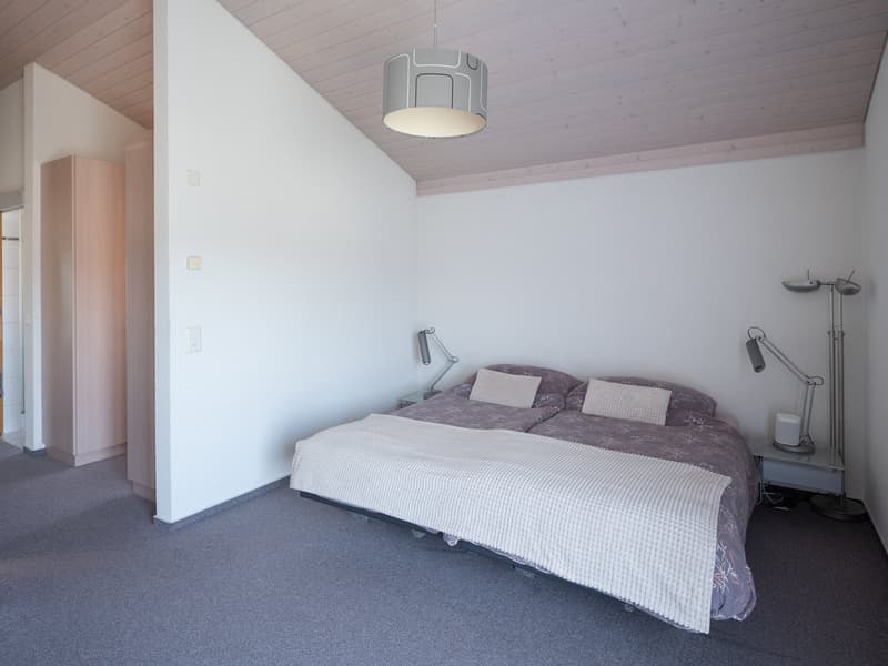 Spacious bedroom with a double bed, white blanket, and two pillows. A wooden wardrobe is on the left, and a nightstand with a lamp is on the right. A ceiling lamp and a floor lamp are also present.