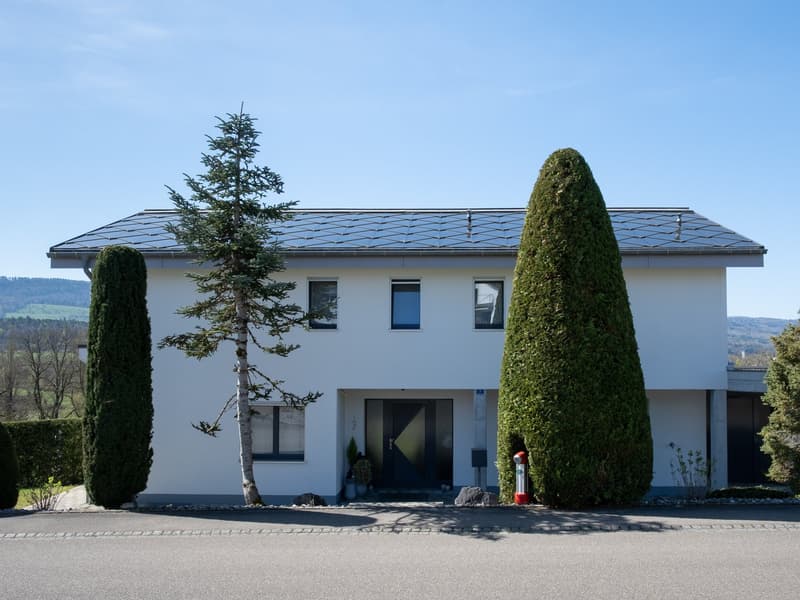 Modern white house with solar panels on the roof, large windows, and a double door entrance.