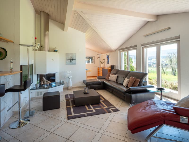 Modern living room with a leather sofa, coffee table, and a fireplace. The room features large windows with a view of the outside. There is a rug on the floor and a side table with a candle. The room is well-lit with a lamp and has shelves with decorative