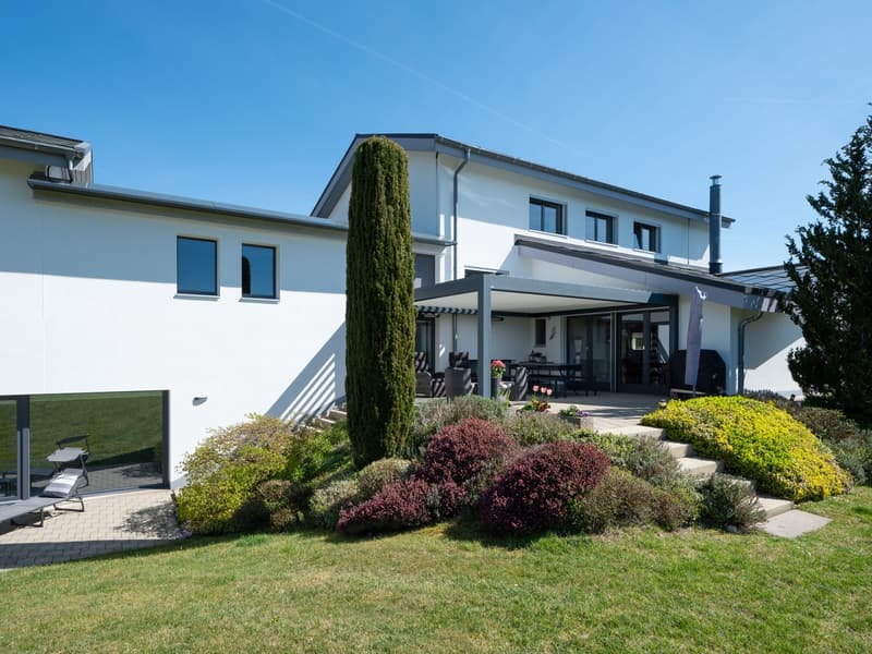 Modern white house with glass windows, a patio with outdoor furniture, and a well-maintained garden.