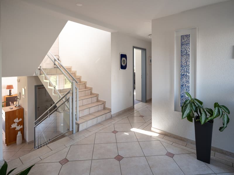 A modern hallway with tiled flooring, a staircase with a glass handrail, and a decorative plant.