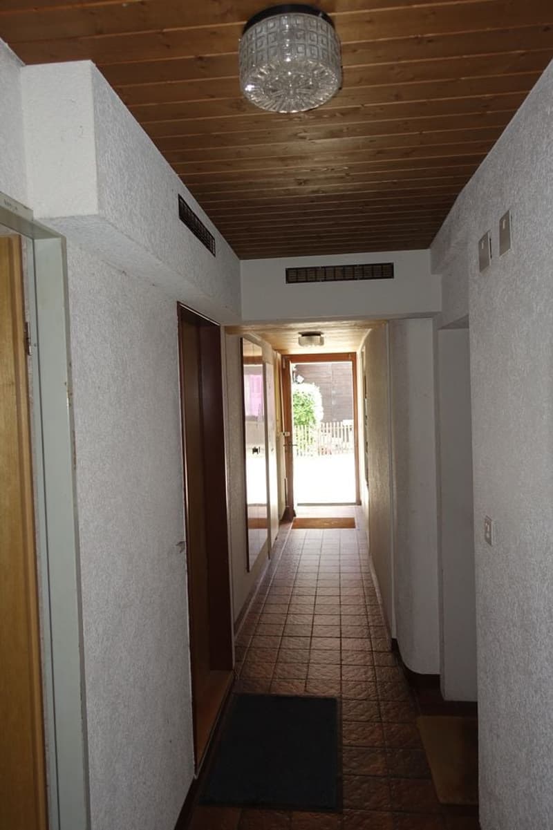 Long hallway with tiled floor, wooden ceiling, doors on either side, open doorway leading to outdoor area.