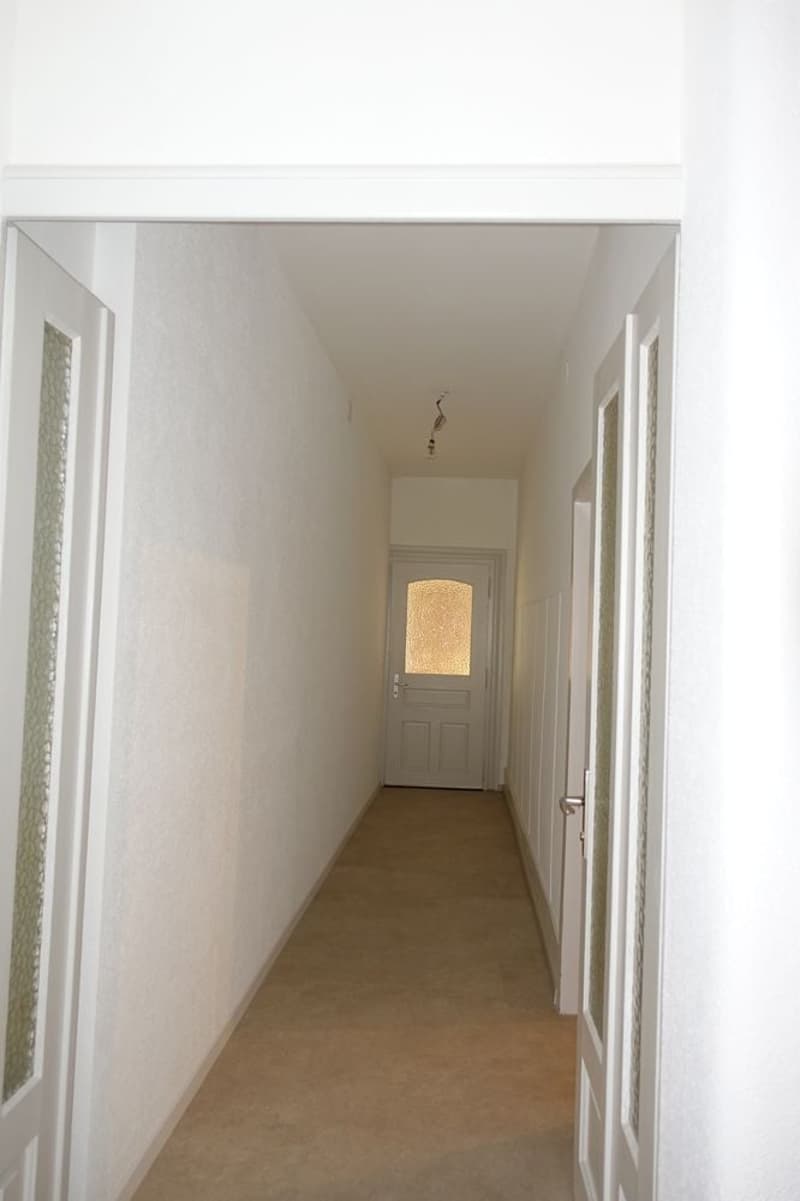 Empty hallway with white walls, doors on both sides, central door, wooden flooring.