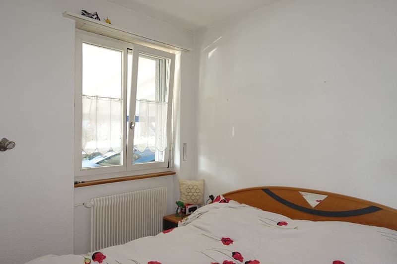 Bedroom with white walls, large window, wooden bed, white floral sheets, nightstand with lamp, white radiator.
