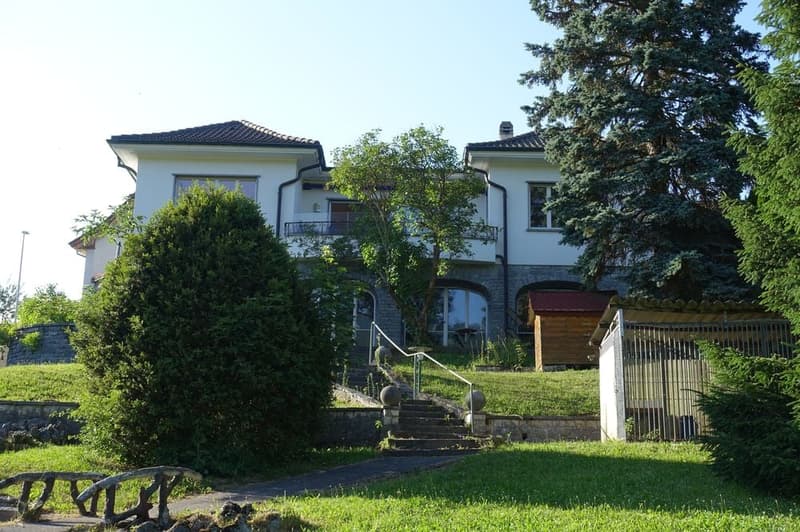2 story house, white exterior, black roof, large balcony, brick foundation, surrounded by trees and shrubs