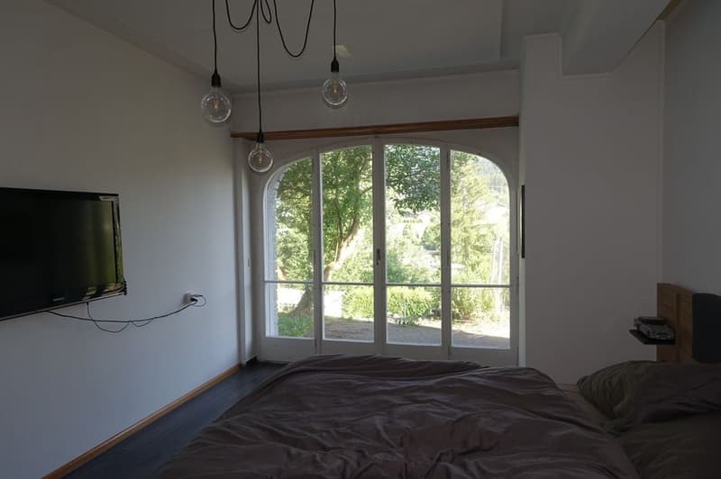 Bedroom with double bed, bedspread, pillows, lamp, TV, wall, windows, hanging plug, large glass doors, garden, trees