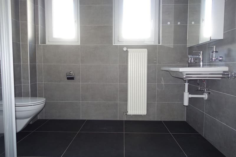 Bathroom with grey tiled walls, black tiled floor, white toilet, white radiator, white sink, white countertop, mirror with lighting, soap dispenser, and faucet