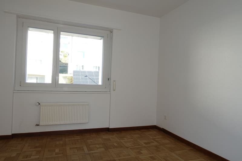 An empty room with white walls, a wooden floor, a window, and a white radiator.