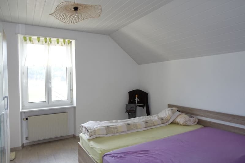 Bedroom with white walls and wooden floor, bed with two sets of covers and pillows, white window with curtains, white radiator, wooden headboard.