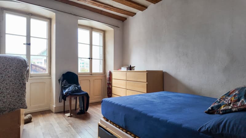 Room with wooden floors, wooden beams on ceiling, two arched windows, wooden chair, blue bed with blue sheets and pillow, pillow with floral print, wooden wardrobe, small table on right of bed.