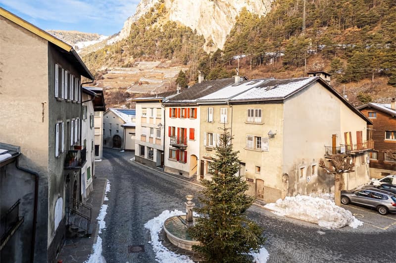The picture shows an aerial view of a small town street lined with buildings and a backdrop of a snow-covered mountain. The street is made of cobblestone, and there is a small fountain in the center with a pine tree. The buildings are a mix of old and new