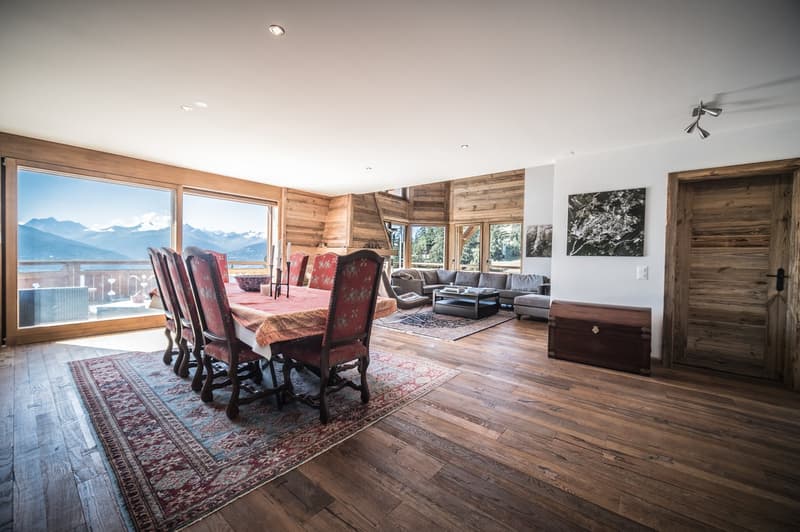 a long dining table with red chairs, a couch, wooden floor, large windows, carpet, wooden chest