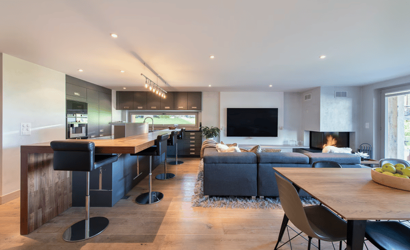Open living space, kitchen island, bar stools, dining table, gray sectional sofa, rug, fireplace, flat screen TV, potted plant