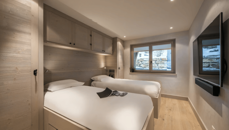 Two beds with white bedding, wooden walls, wooden floors, window with a view of a stone wall, mounted television, cabinets with drawers, night lamps on each bed