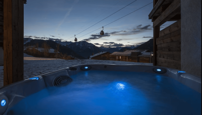 Outdoor hot tub with blue light, snowy mountains, ski lifts in the distance, twilight sky