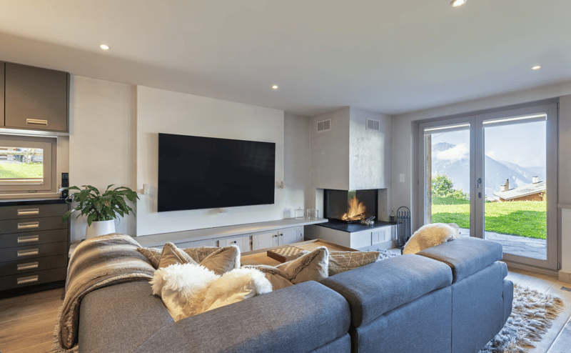gray sectional sofa, large windows, fireplace, TV, potted plant, rug, pillows, wooden floor, kitchen cabinets, chest drawers