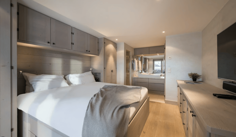 A bedroom with a double bed and white linens, gray throw, white nightstand, wooden dresser with TV and remote, built-in storage, and a glimpse of a bathroom with a sink.
