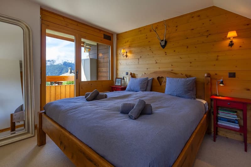 Bedroom with wooden walls, queen size bed, large glass door, balcony view, mountain view, night stand, lamp, mirror, deer head decoration, wooden bed frame, two nightstands with drawers and stack of books, blue linens