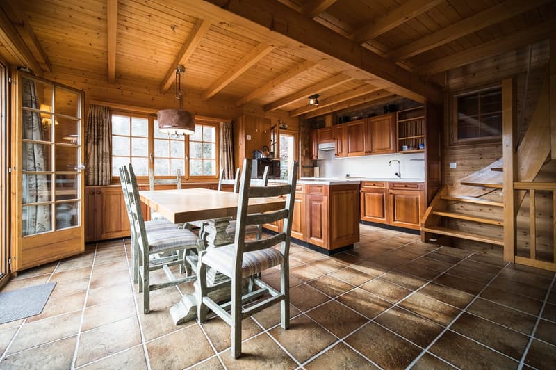 Spacious open-plan kitchen and dining area with wooden ceiling beams, large windows, wooden cabinetry, tile flooring, and a dining table with chairs