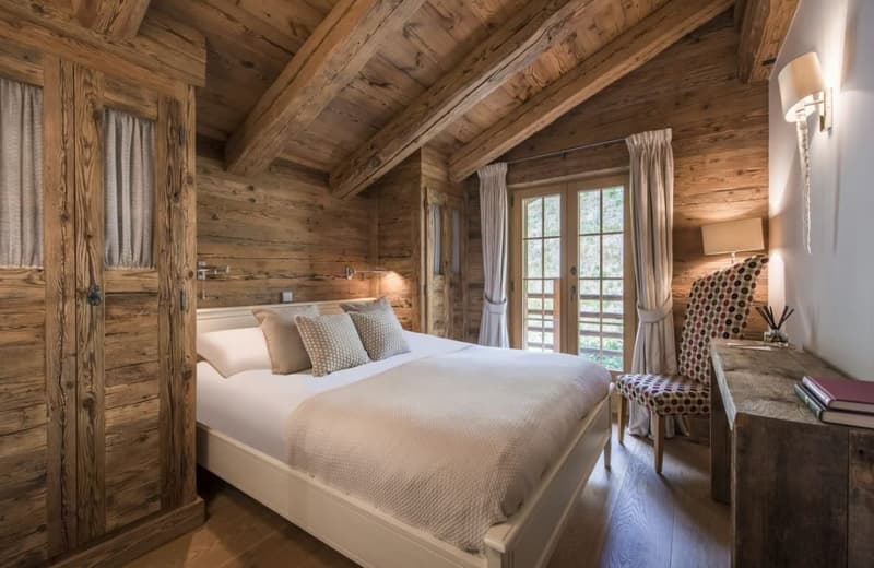 Rustic wooden cabin bedroom with a large bed, wooden walls and ceiling, curtains, and a balcony or terrace visible through the window.
